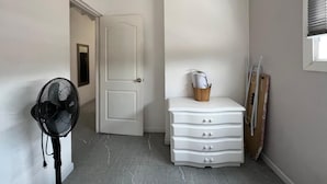Un couloir avec une porte blanche, une commode blanche à tiroirs, un ventilateur et une fenêtre avec des stores.