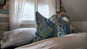 A bedroom with a bed, floral-patterned pillows, a white pillow, a window with sheer curtains, and a mirror on the wall. - Cozy Country Cottage in the heart of Skagit Valley! (Mount Vernon)