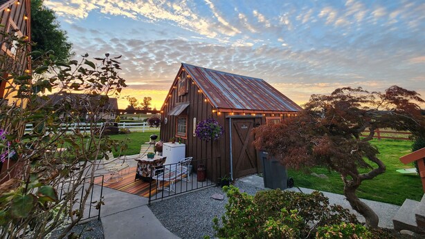 A rustic barn with a sloped metal roof, surrounded by a well-maintained garden and a wooden fence. - Cozy Country Cottage in the heart of Skagit Valley! (Mount Vernon)