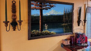 A framed landscape painting of a lake with mountains and trees, a wooden shelf with candles and a decorative pot, and a window with blinds. - Cozy Country Cottage in the heart of Skagit Valley! (Mount Vernon)