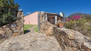 A stone-walled entrance with a number sign, a building with a pink facade, and a clear blue sky. - Terraced house with swimming pool (Tertenia)