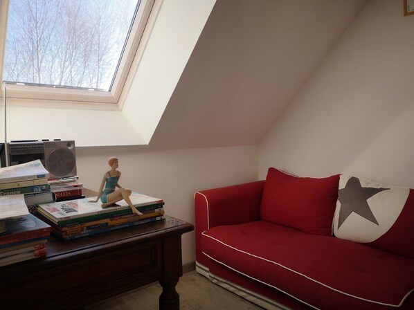 A room with a sloped ceiling, a red sofa, a small table with books and a figurine, and a window with a view of bare trees.