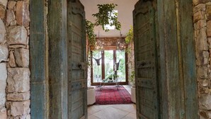 Un pasillo con paredes de piedra rústica y puertas metálicas verdes que dan a una habitación con alfombra roja, un candelabro y vistas a la vegetación exterior.