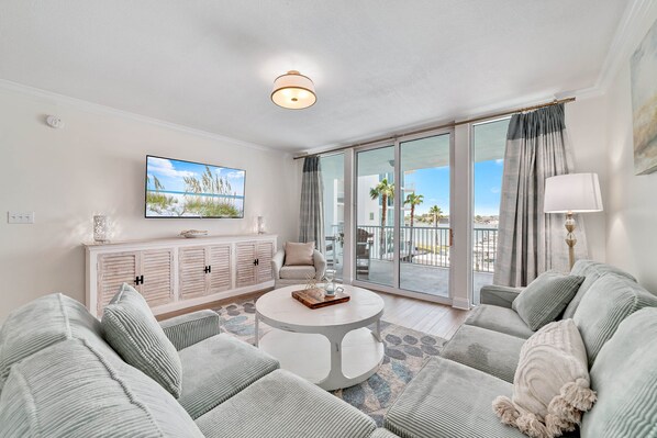 A modern living room with a round white coffee table, grey sofas, a flat-screen TV, a white cabinet, and a balcony with a view of palm trees.