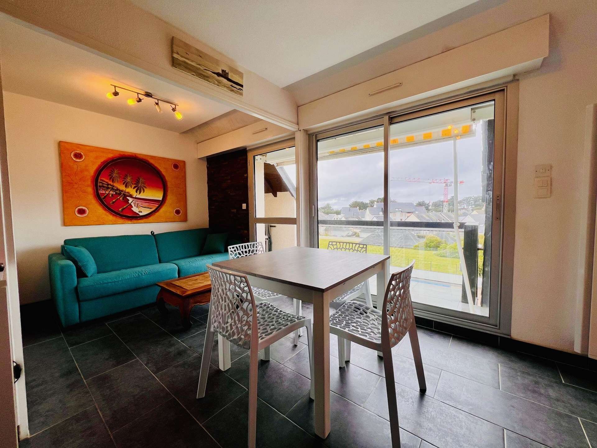 A modern living room with a teal sofa, a dining table with chairs, and a large window offering a view of the outside.