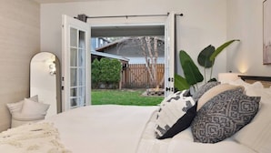 A bedroom with a large bed, a mirror, and a view of a backyard through an open door. - Sanders Beach Retreat, walk to DT & Sanders beach! (Coeur d'Alene)