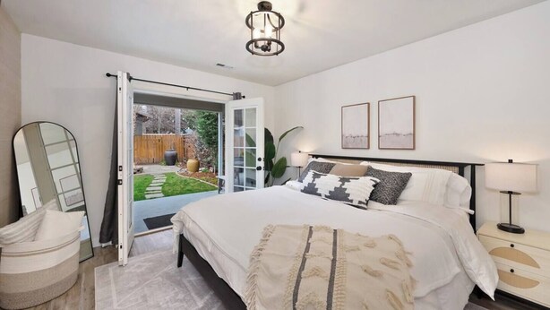 A bedroom with a large bed, a bedside table with a lamp, a mirror, and a view of a garden through an open door. - Sanders Beach Retreat, walk to DT & Sanders beach! (Coeur d'Alene)