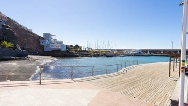 A coastal promenade with a wooden deck, a rocky cliff, and a marina with boats.