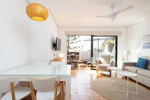 A modern living room with a glass dining table, white sofas, a ceiling fan, and a view to the outside.