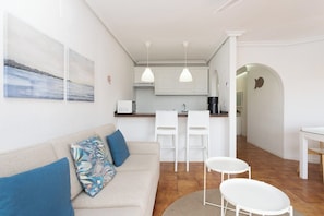 Et moderne interiør med en sofa, to stole, et lille bord og et køkkenområde med hvide skabe og hængende lamper.