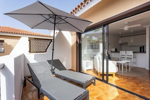 A balcony with two sun loungers and a large umbrella, a sliding glass door leading to a kitchen area with white furniture.