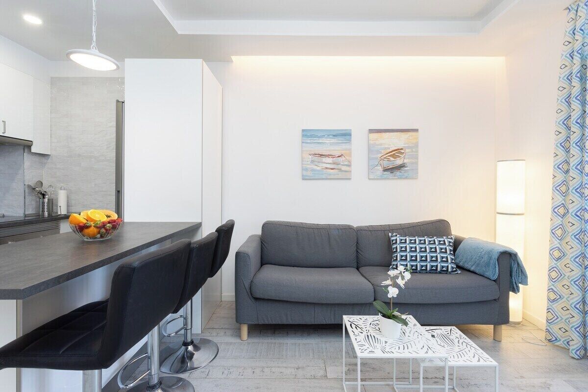 En moderne stue med en grå sofa, et hvidt sofabord og et spiseområde med en sort stol og en skål med frugt.