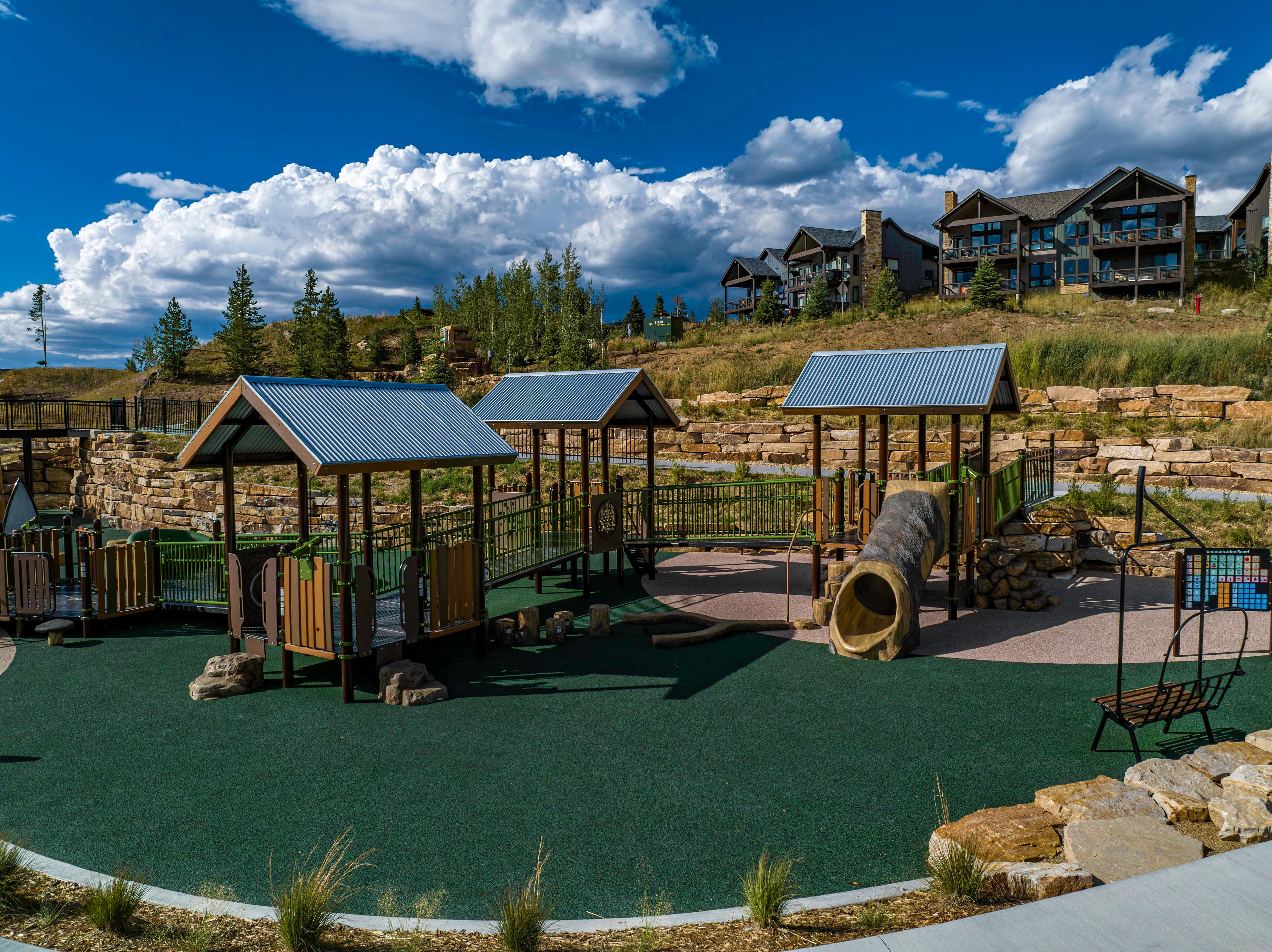 A playground with climbing structures and a slide, surrounded by a landscaped area with a view of residential buildings.