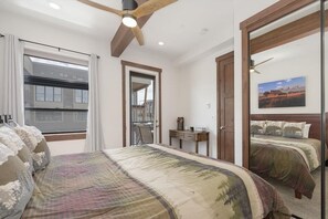 A bedroom with a large bed, a ceiling fan, a window with curtains, a door, and a small table with a vase.