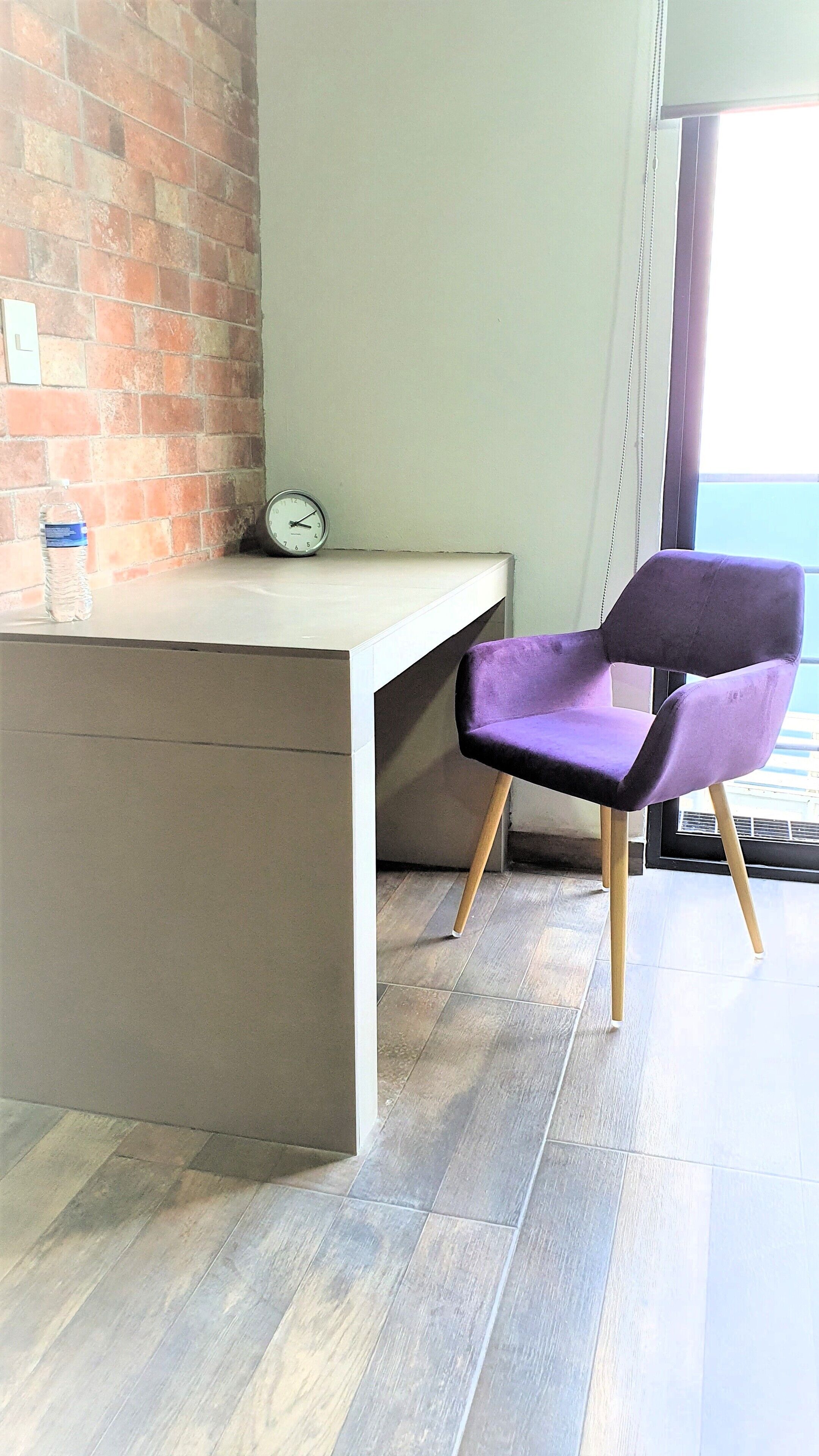 Habitació moderna amb paret de maó, escriptori blanc, cadira morada i rellotge.