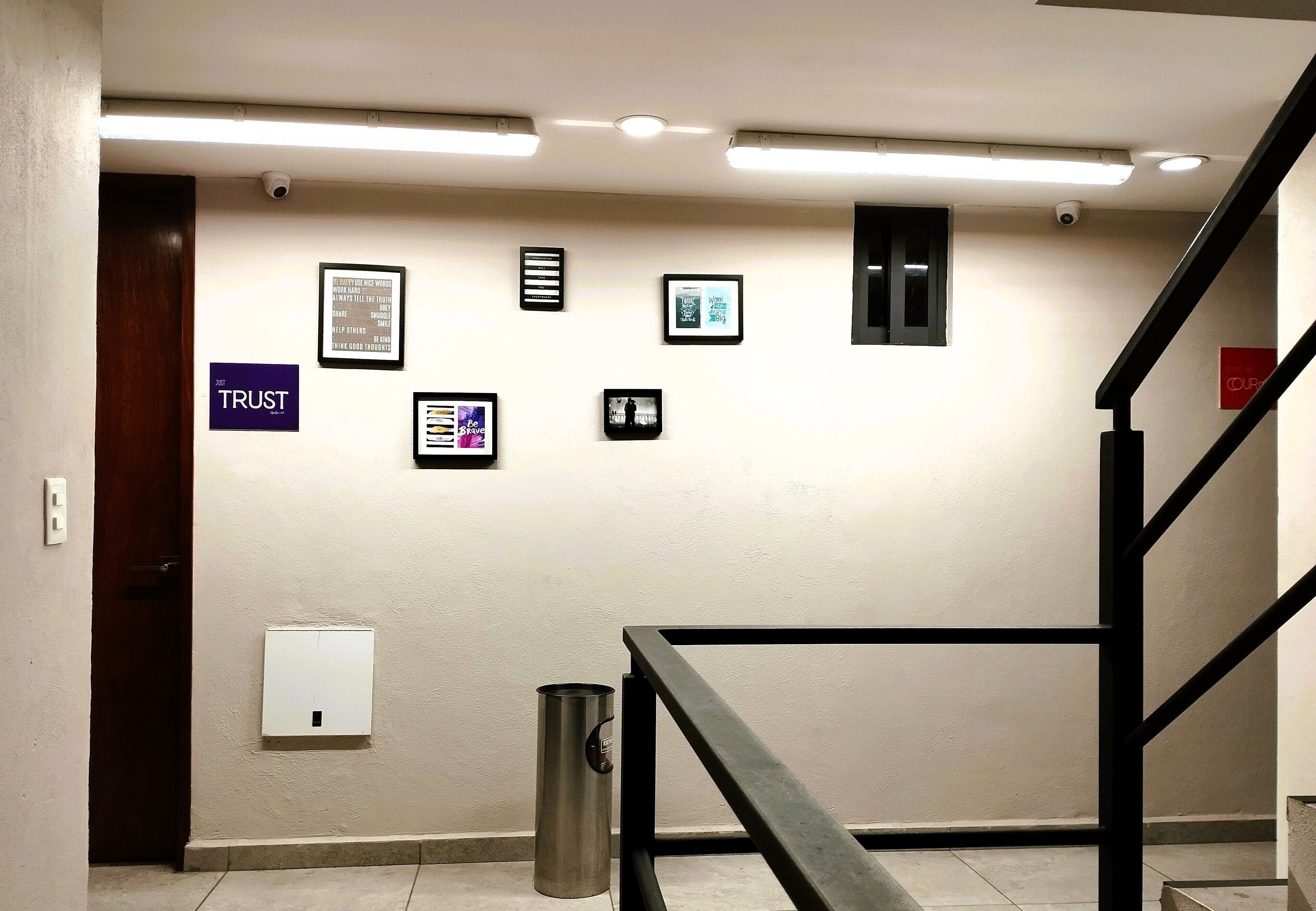 Un pasillo con una escalera, un cubo de basura sujeto a la pared y varias fotografías enmarcadas.