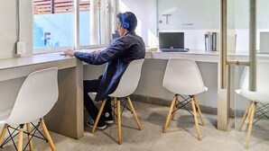 Una persona sentada en un escritorio con una computadora portátil, en una habitación con sillas blancas y un ventanal.