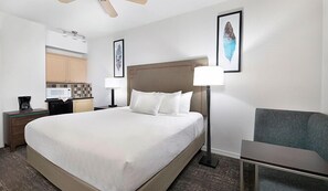 Habitación de hotel con una cama grande, un escritorio con lámpara, una silla y una kitchenette al fondo.