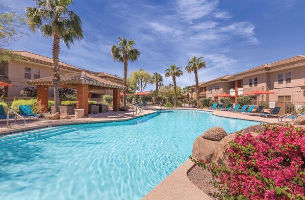 A swimming pool with lounge chairs and palm trees, surrounded by buildings with a tiled roof and a clear sky.
