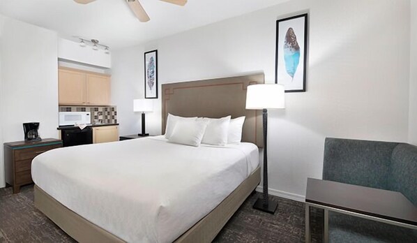 A hotel room with a large bed, a desk with a lamp, a chair, and a kitchenette in the background.