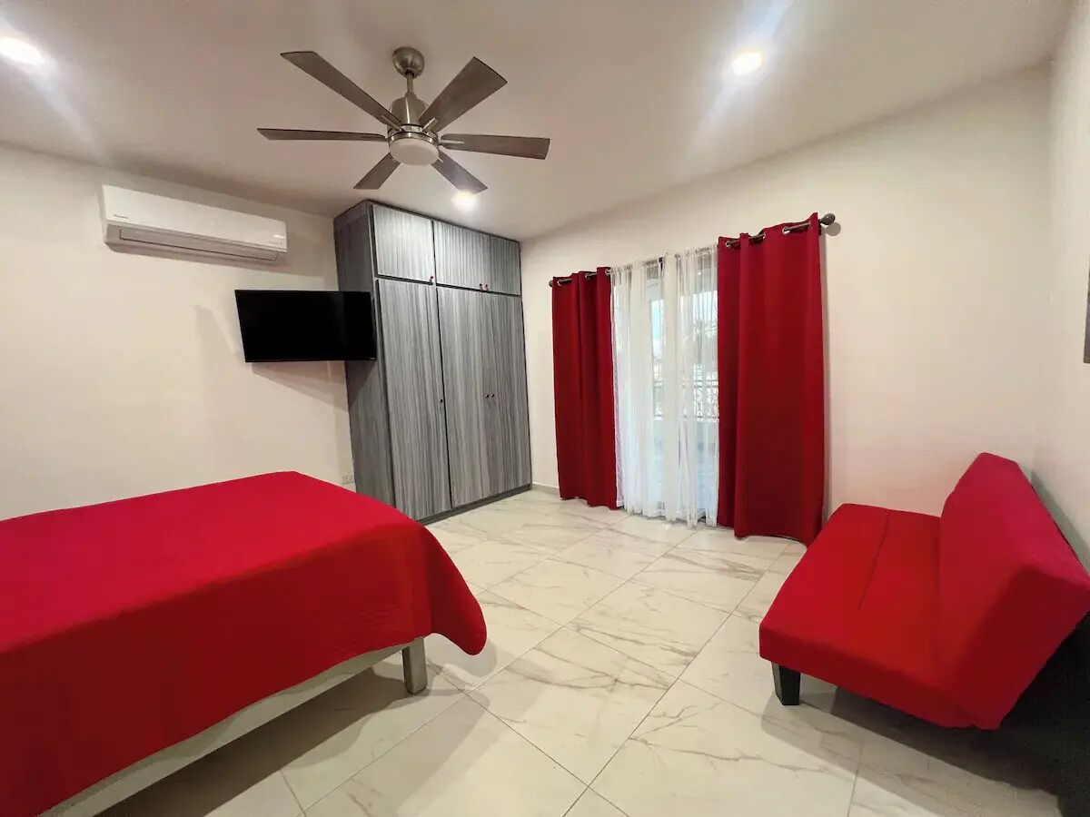 Una habitación con dos camas individuales, ventilador de techo, aire acondicionado, televisión y ventana con cortinas rojas.