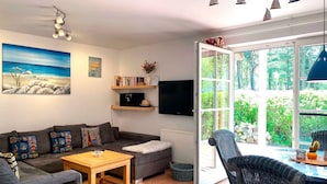 Una sala de estar moderna con un sofá gris, una mesa de centro de madera y una silla de mimbre. Hay un televisor en la pared, una estantería y un gran ventanal con vistas a la vegetación.