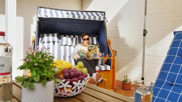 A person reading a book in a cozy, striped chair with a basket of fruit and a bottle on a table.