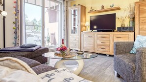 Ett mysigt vardagsrum med en soffa, en fåtölj och ett soffbord i glas. Det finns en tv på ett träskåp och ett stort fönster med gardiner.