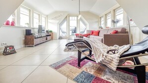 Un amplio loft con techo alto, grandes ventanales, una zona de estar acogedora con un sofá y sillón, una pequeña mesa de comedor y un televisor sobre un mueble de madera.
