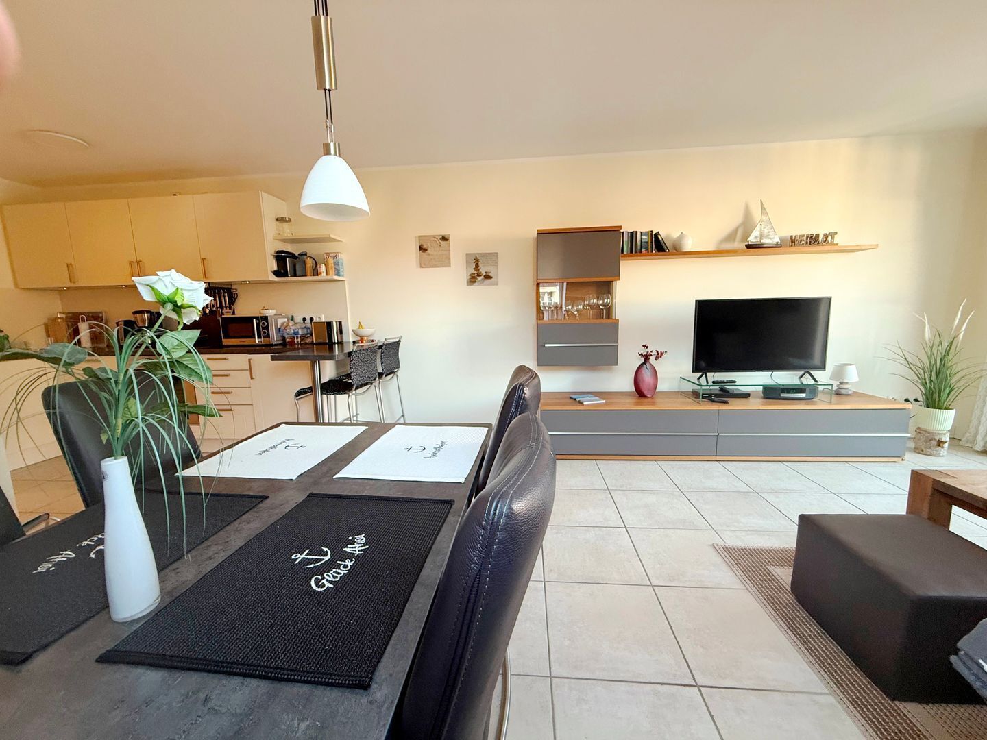 A modern living room with a dining area, a kitchen in the background, and a TV on a stand.