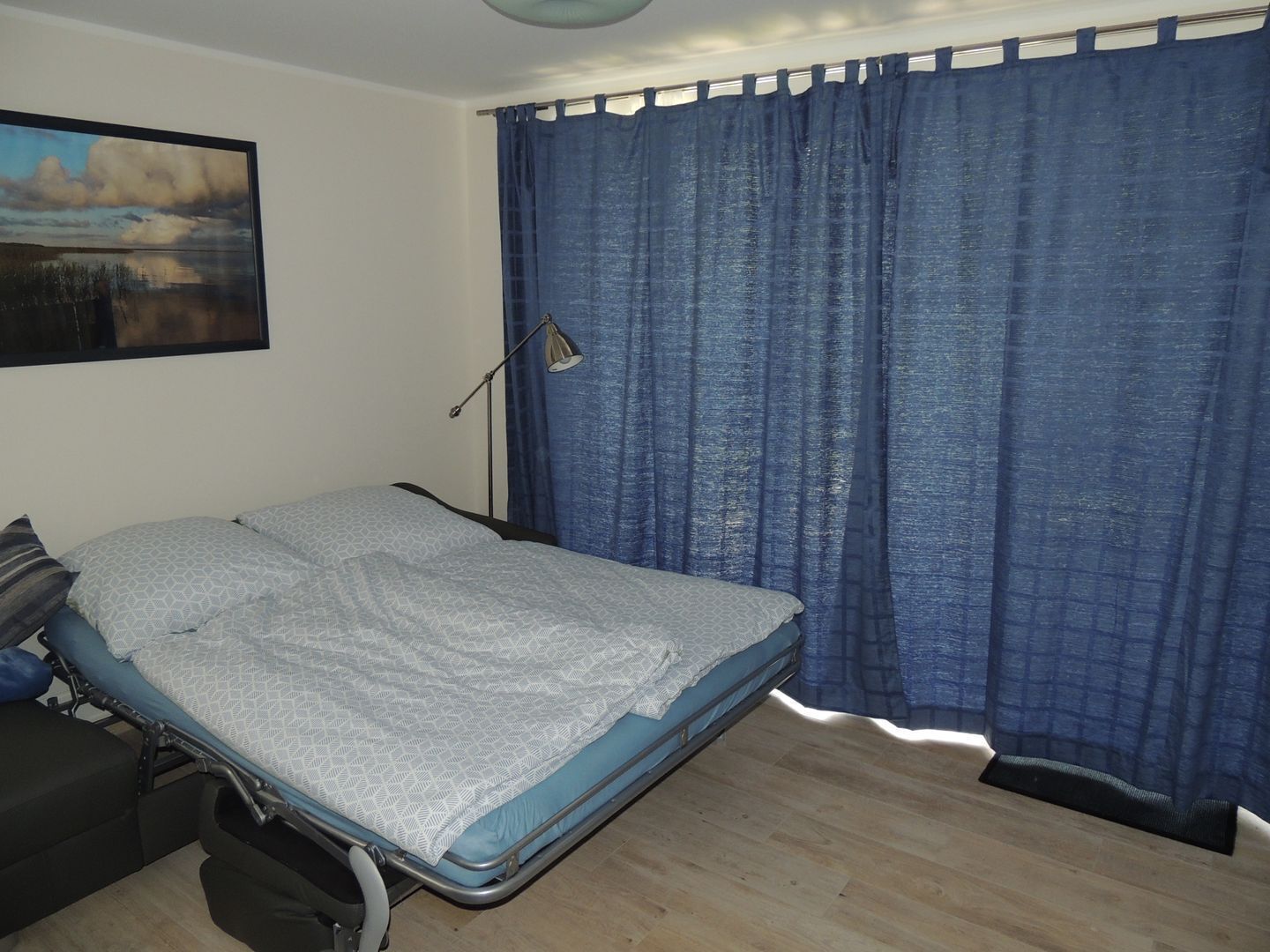 Een enkel bed met metalen frame, een blauw gordijn en een schilderij aan de muur.