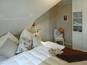 Un dormitorio con cama, almohadas, mesita de noche, una silla, estante con adornos y armario.