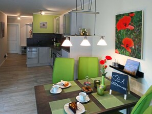Ein moderner Essbereich mit einem Tisch für zwei Personen, dekoriert mit einer Torte und einer Tasse. Ein grüner Stuhl und eine Vase mit roten Blumen sind zu sehen. An der Wand ist eine Beschilderung erkennbar.