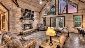A log cabin interior with a stone fireplace, leather sofas, and large windows offering views of the outdoors.