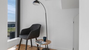 A modern room with a high ceiling, a floor lamp, a grey chair, a small round table with a magazine, and a window with a view of buildings. - Ocean front home in Skagen with WiFi (Skagen)