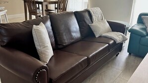 A brown leather sofa with white cushions, a dining table with chairs, and a blue chair.