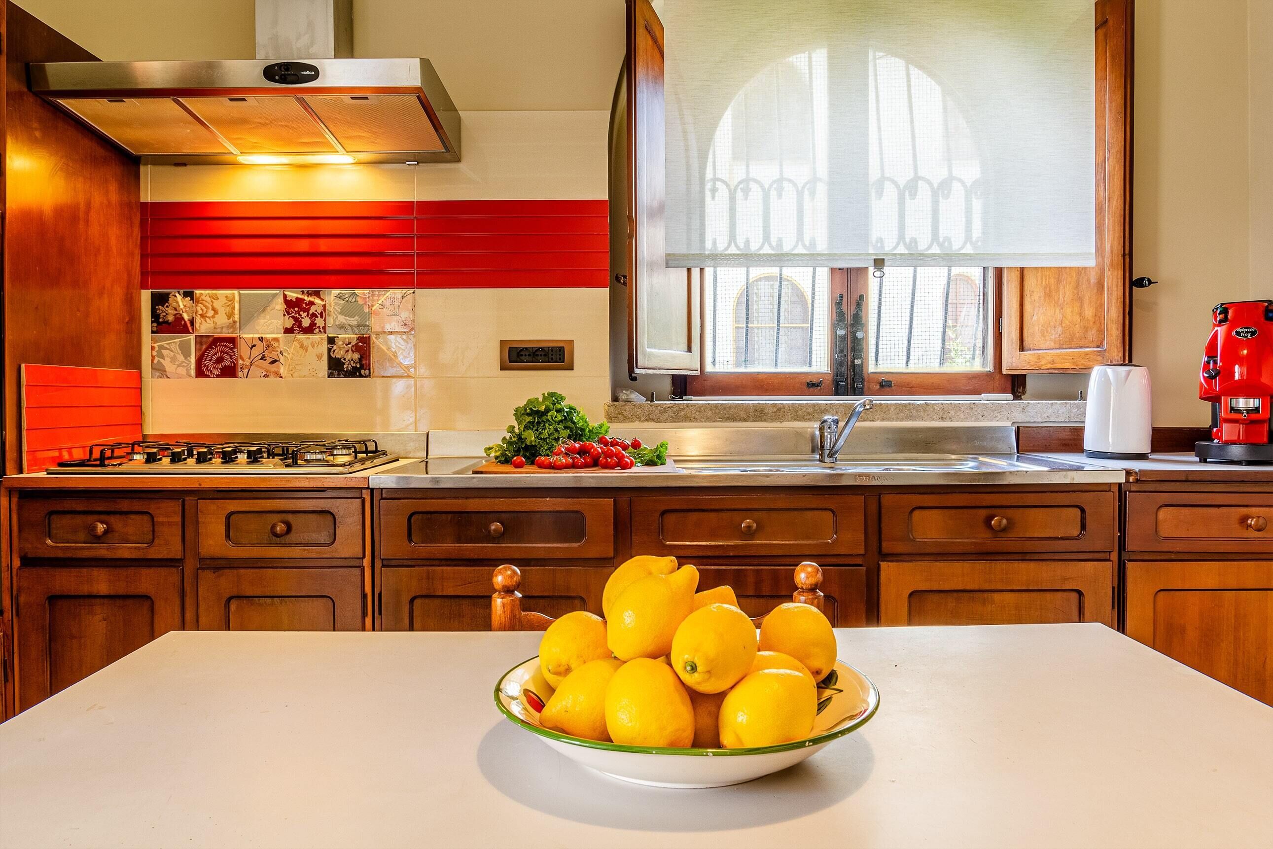 A kitchen with a bowl of lemons on the counter, wooden cabinets, a stove, and a window with a view.