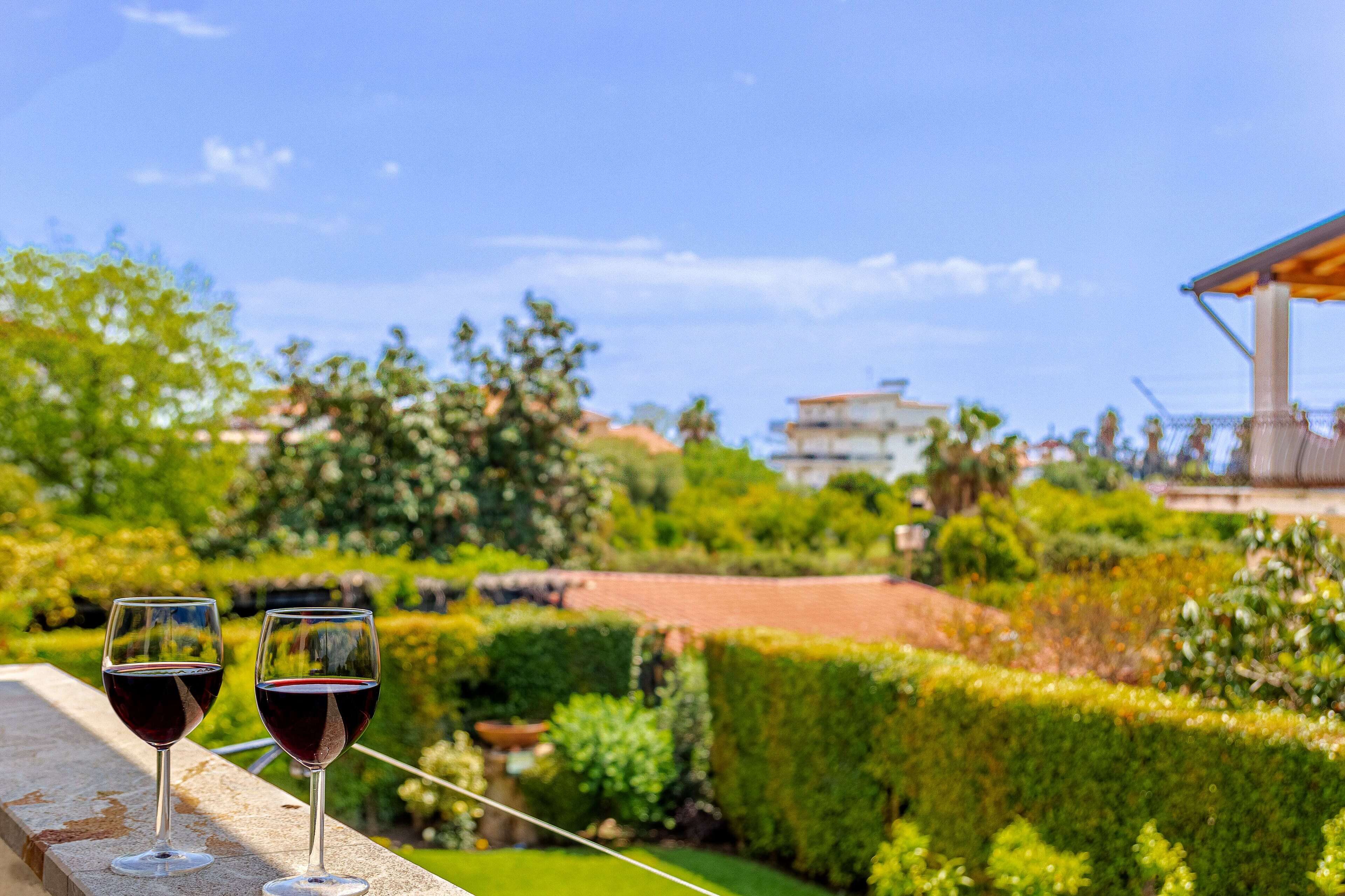 Two wine glasses on a balcony with a view of a well-maintained garden and a building in the background.