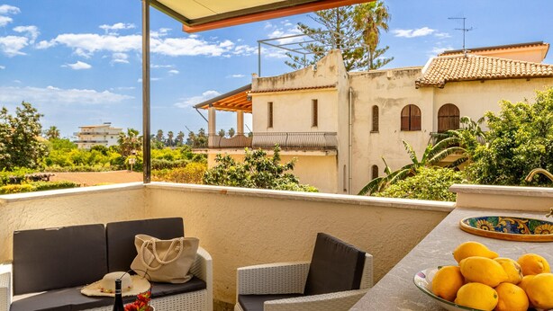 A balcony with wicker furniture, a table with a bottle and glasses, and a bowl of lemons. - Villa Magnolia - Apartment in villa (Giardini Naxos)