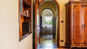 A hallway with a wooden cabinet, a bookshelf, and an arched doorway leading to a room with a sofa and a view of greenery outside. - Villa Magnolia - Apartment in villa (Giardini Naxos)