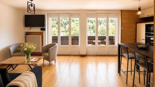 En moderne stue med sofa, spisebord, TV og et køkkenområde med barstole.
