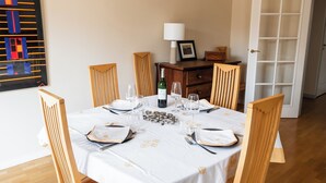 Mesa de comedor para seis personas, con manteles blancos y detalles dorados, sillas de madera, una botella de vino y una lámpara sobre un mueble de madera.