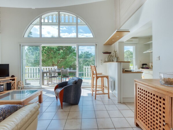 A spacious living area with a large arched window, a dining table set for four, a sofa, and a kitchen area with a wooden cabinet and a bowl on the counter.
