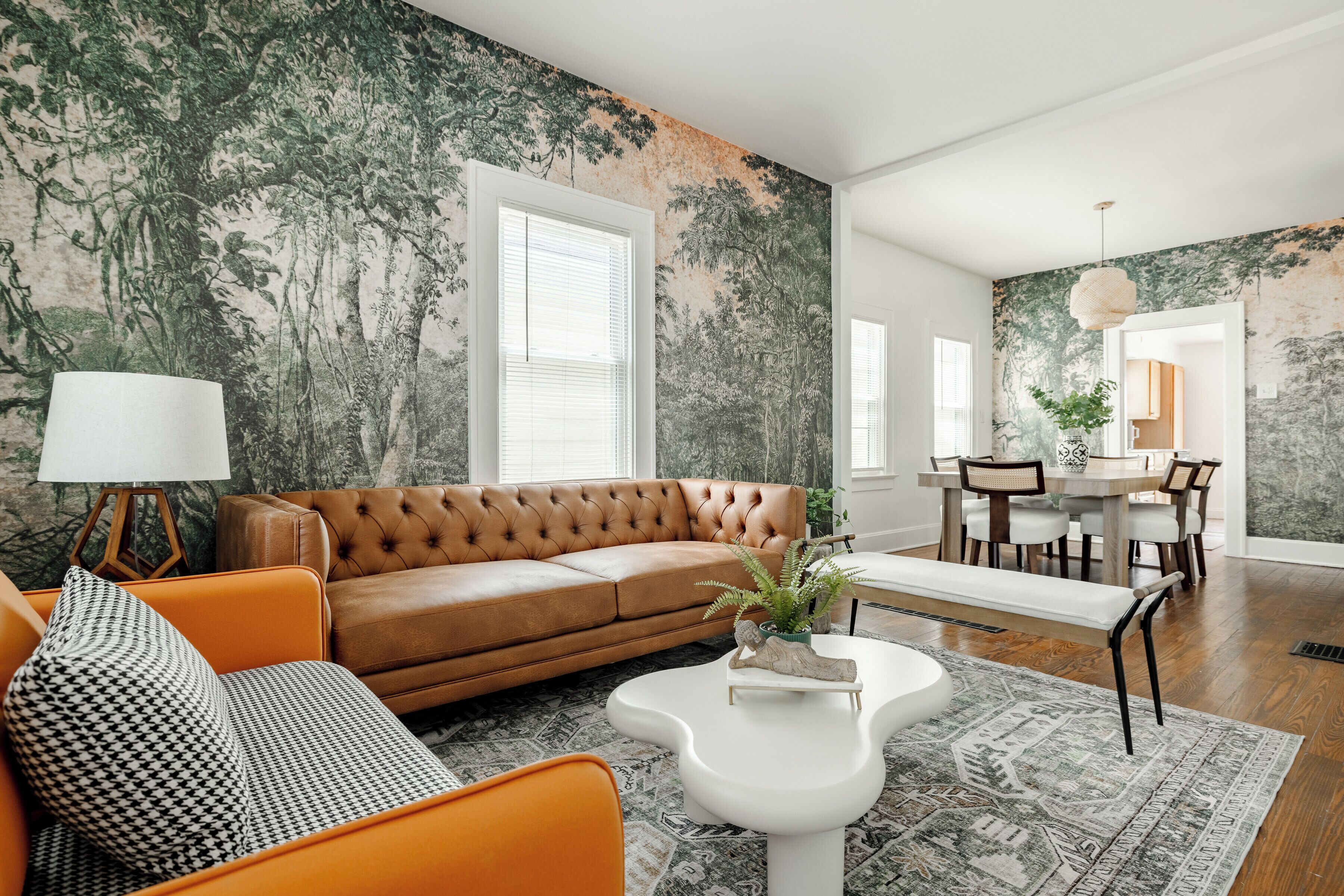 Een woonkamer met een bruine leren bank, een oranje fauteuil, een witte salontafel en een kamerkleed met een patroon.