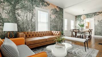 Een woonkamer met een bruine leren bank, een oranje fauteuil, een witte salontafel en een kamerkleed met een patroon.