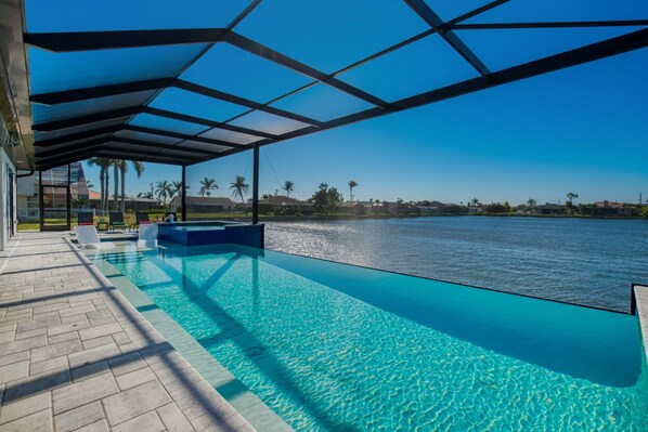 A swimming pool with a clear view of the water, a covered area with chairs, and a distant view of palm trees and buildings. - VILLA STINGRAY – HEATED POOL & SPA | LAKE VIEW | BOAT RENTAL + TOURS VOUCHER INCL. (Cape Coral)