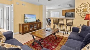 A living room with a sofa, a coffee table, a TV, and a ceiling fan. There is a kitchen area with a bar and chairs. - Lake View Pool Home With Game Room (Haines City)