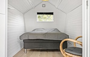 Une pièce de taille modeste, aux murs recouverts de panneaux blancs, comprenant un lit, une fenêtre et une chaise en osier.