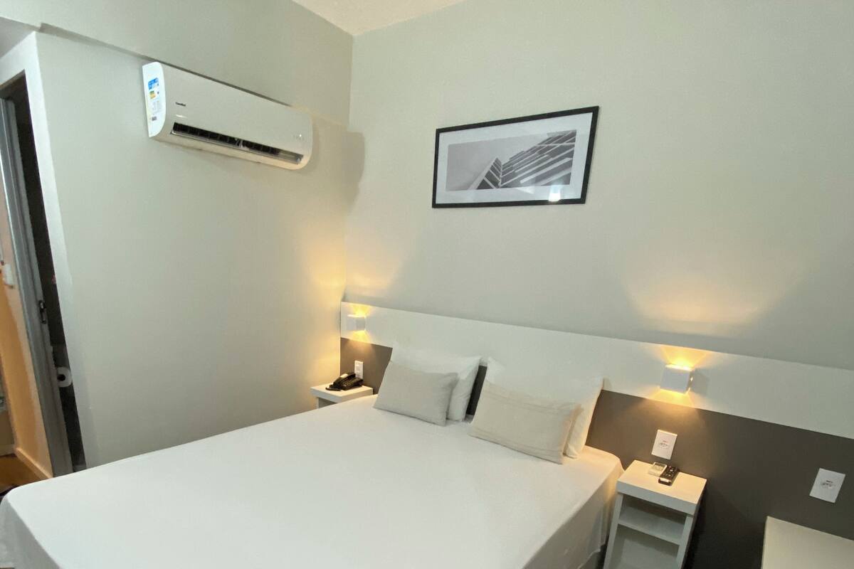 Habitació d'hotel amb un llit gran, taules de nit amb làmpades, aire condicionat fixat a la paret i un quadre al mur.