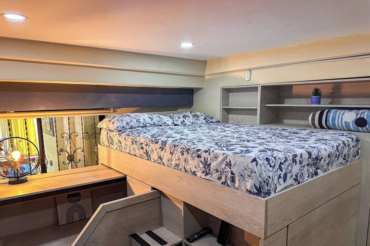 Una cama litera con una colcha estampada, un escritorio con una lámpara, y una estantería con objetos decorativos.